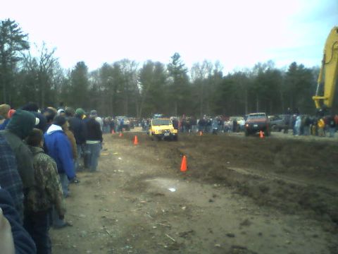 1976 GMC C/K Pickup - A crowd watches as off-road vehicles navigate a muddy track, with a large yellow excavator visible in the background.