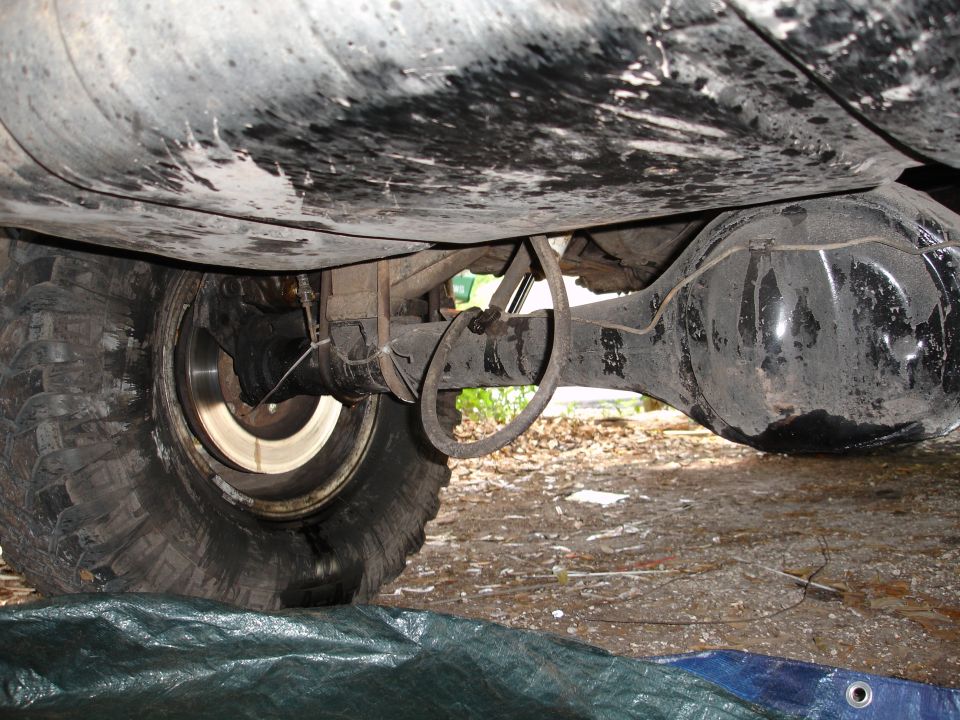 Underside view of a vehicle's rear axle, differential, leaf springs, and tire. A green tarp is below the tire