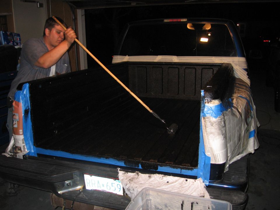 Man rolling black bed liner into a blue pickup truck bed masked with tape and newspaper at night