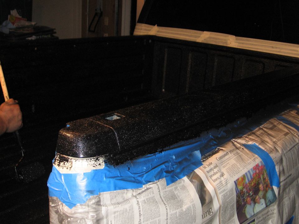 Applying black textured coating to a truck's tailgate cap with a roller. Blue tape and newspaper protect the surrounding area