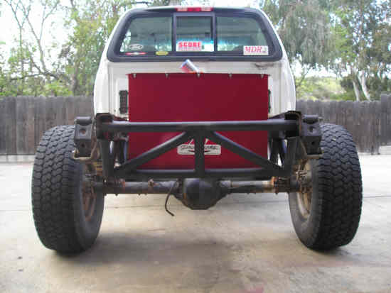 Rear view of a white Ford pickup truck chassis with a red panel and custom roll cage. Tires are large and knobby