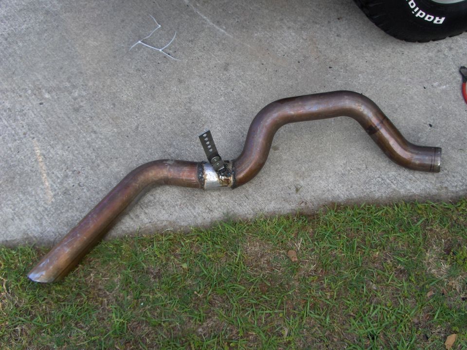 Bent exhaust tailpipe with welded hanger strap lying on concrete next to grass