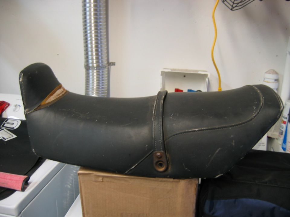 Black motorcycle seat with a leather strap and metal buckle, resting on a cardboard box