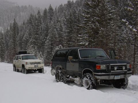1990 Ford Bronco - Two SUVs navigate a snowy forest road, with the Ford Bronco in the foreground and a Jeep Grand Cherokee behind it.