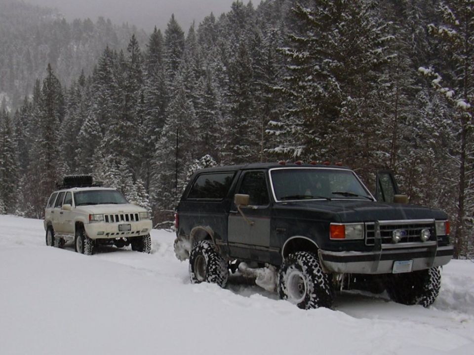 Black Ford Bronco with off-road tires in snow, front 3/4 view. A white Jeep Grand Cherokee is behind it