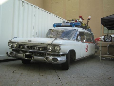 1990 Ford Bronco - white Cadillac ambulance-style car fitted with roof-mounted equipment, lights, and a Ghostbusters logo, parked beside a shipping container.