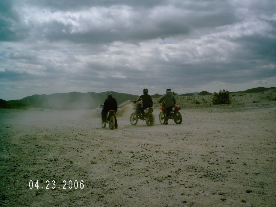 Three dirt bike riders stopped on a dusty desert trail under cloudy skies