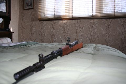 2004 Jeep TJ - A rifle rests on a bed in a room with blinds and patterned wallpaper.