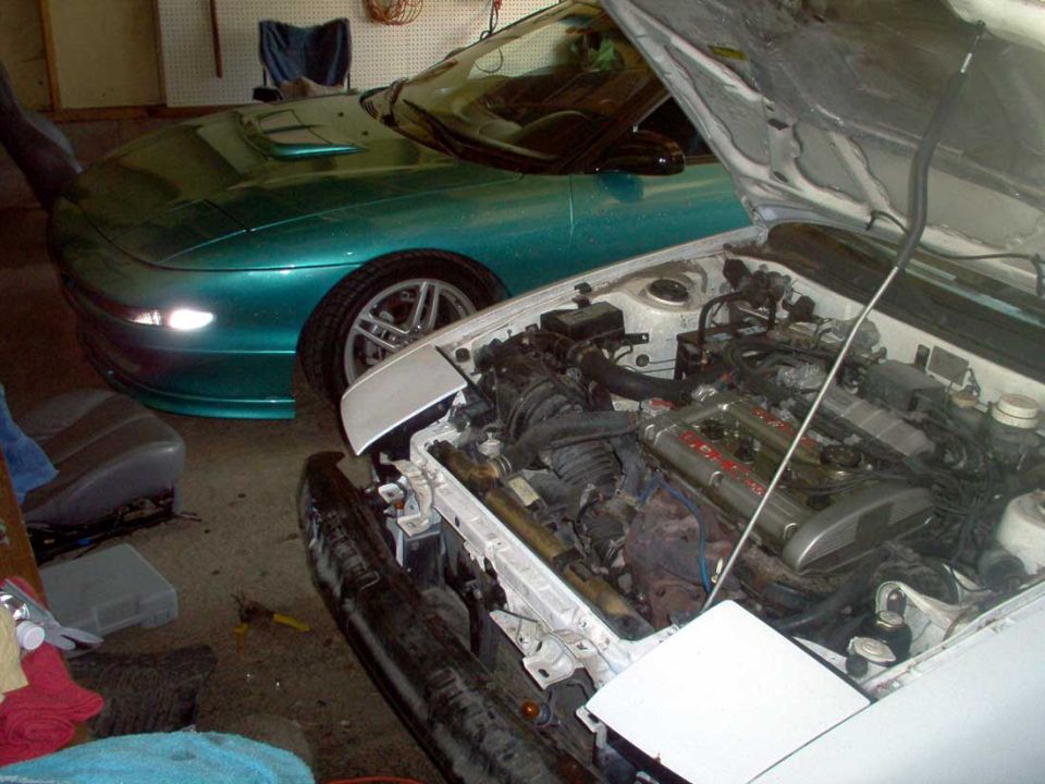 White car with hood up showing engine bay, teal coupe parked beside it in a cluttered garage.