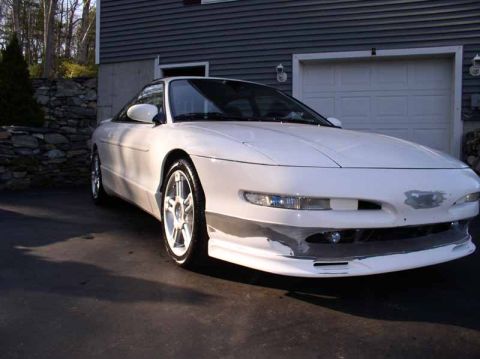 1996 Ford Probe - A white coupe with an aggressive body kit is parked in front of a garage, showing damage to its front bumper.