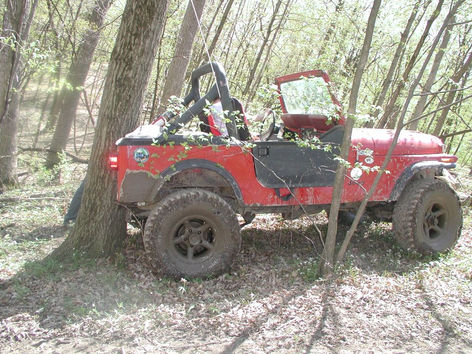 Red Jeep Wrangler with black doors and roll cage, partially covered in leaves, parked near a tree in a wooded area