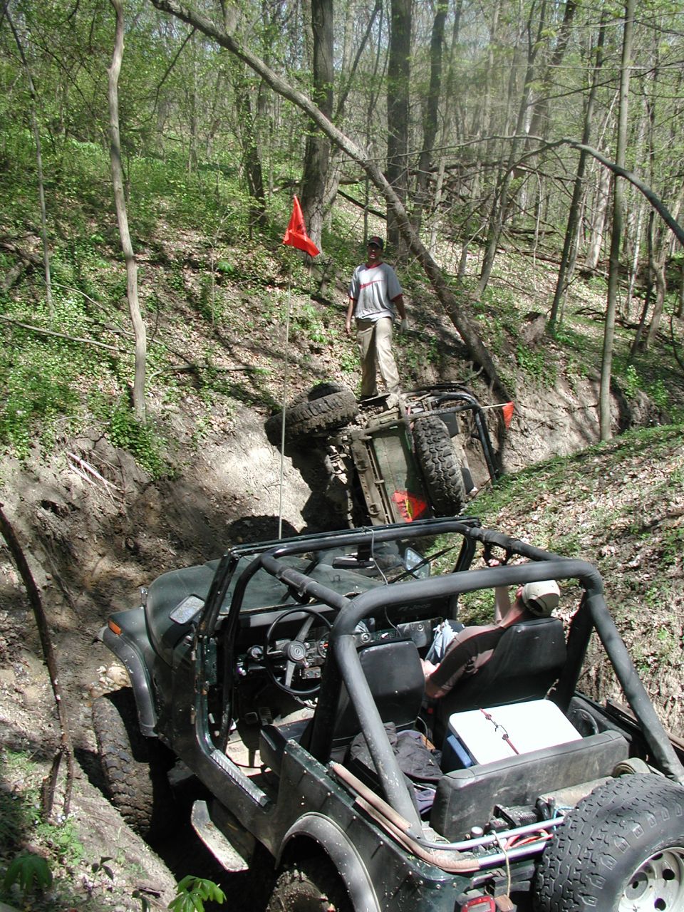 Two off-road vehicles in a wooded ditch, one on its side with a person standing on it