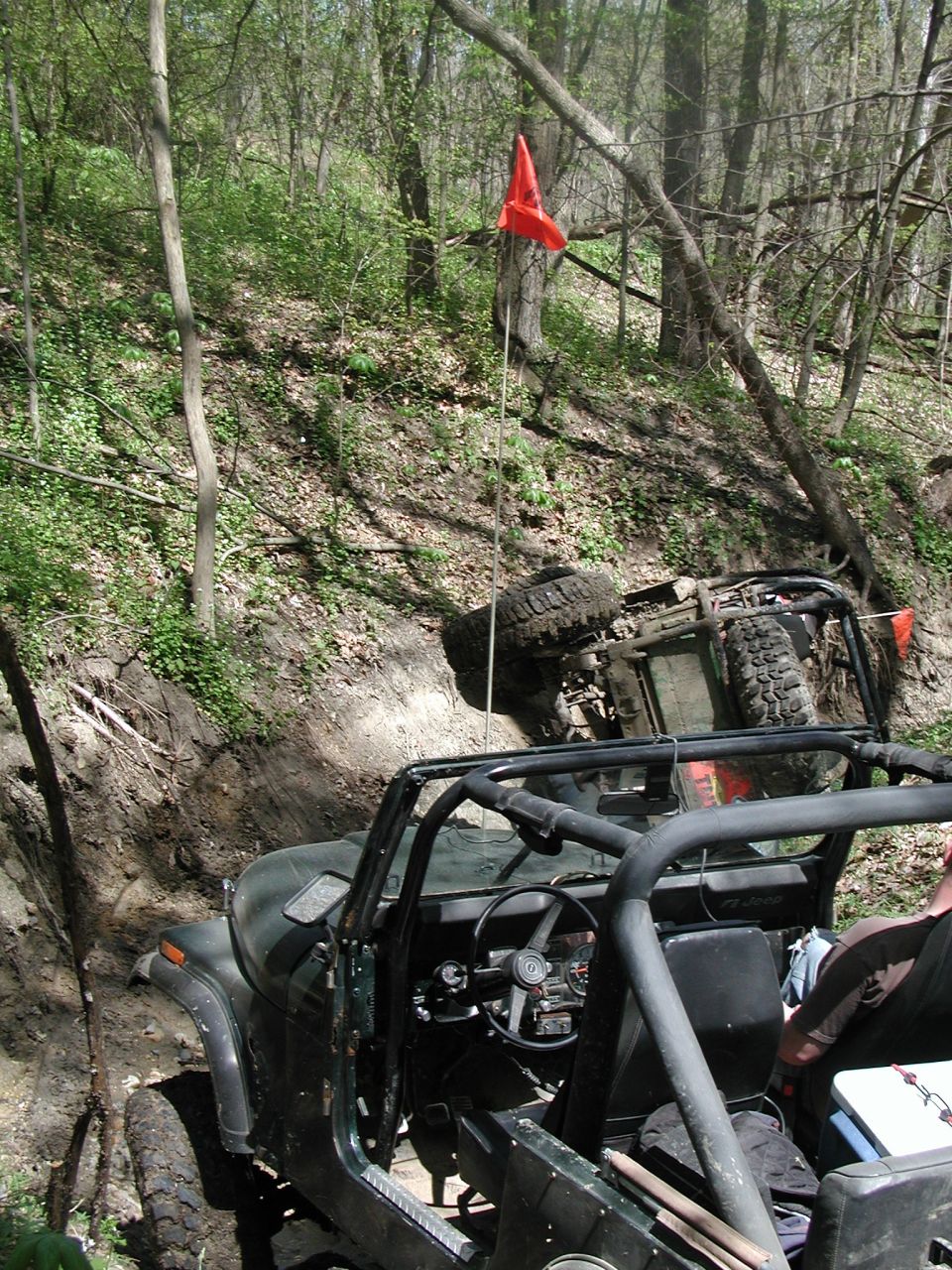 Two off-road vehicles on a muddy trail, one is overturned on its side. A red flag is visible on a pole