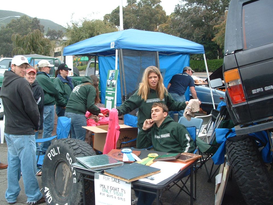 People gathered around a table and tent at an outdoor event, with a large tire and part of a black SUV visible