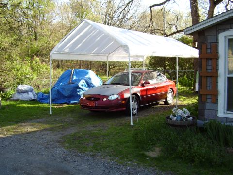 1997 Ford Taurus - A red sedan is parked under a temporary carport, with a blue tarp-covered object behind it.