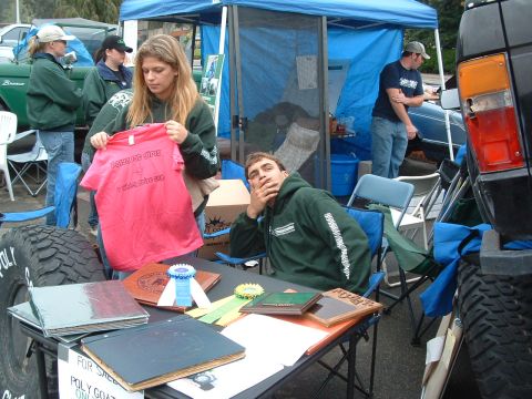 1994 Jeep YJ - An outdoor event with people gathered around a table displaying merchandise and awards, with vehicles visible in the background.