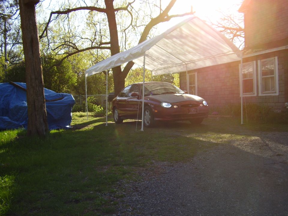 1997 Ford Taurus parked under a white carport next to a house. Another vehicle is covered with a blue tarp to the left