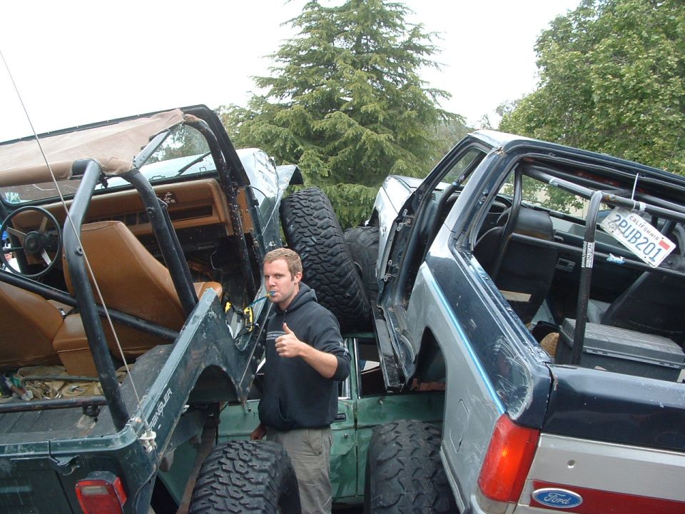 Two Jeep YJs, one green and one dark blue, parked close together. A man stands between them giving a thumbs up