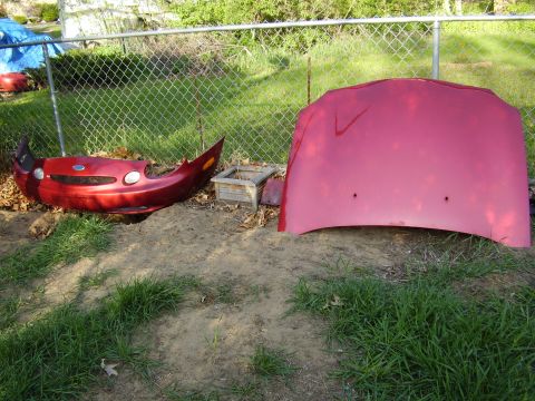 1997 Ford Taurus - A red 1997 Ford Taurus bumper and hood are displayed outdoors, separated and resting on the ground.