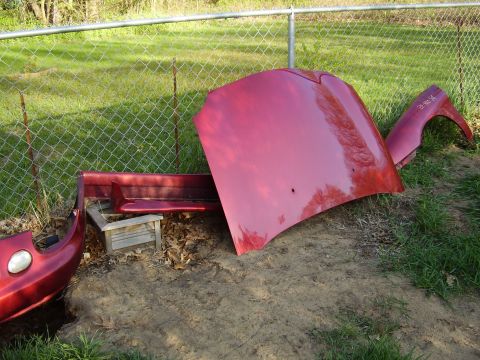 1997 Ford Taurus - Disassembled red car parts, including a hood and fender, are laid out on the ground near a fence.