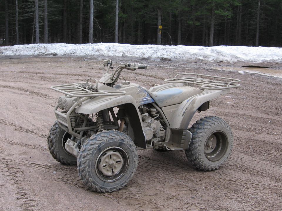 Mud-covered gray Yamaha ATV parked on dirt with snow and forest behind