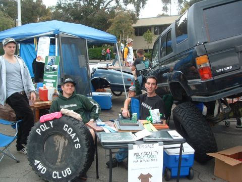 1994 Jeep YJ - A lifted black SUV is parked next to a table with people and a large tire with club text.
