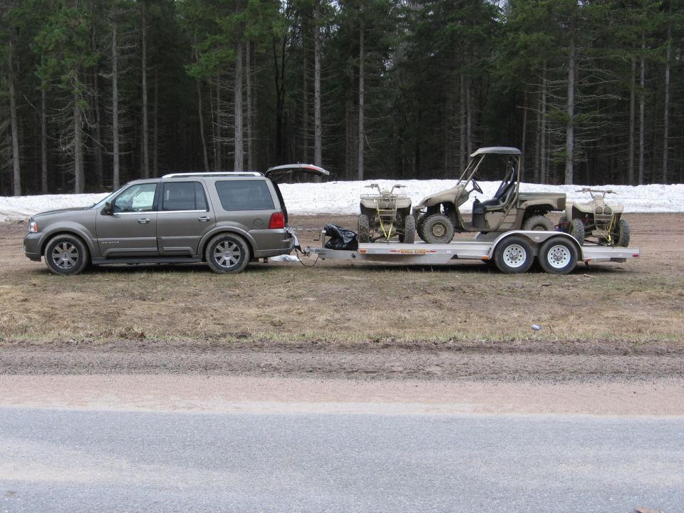 Gold Lincoln Navigator SUV towing trailer with ATVs and a UTV roadside near forest