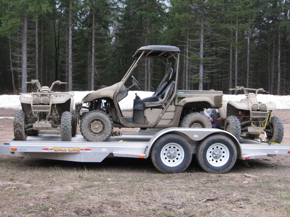 Muddy tan Yamaha UTV and two ATVs loaded on an aluminum trailer outdoors near trees.