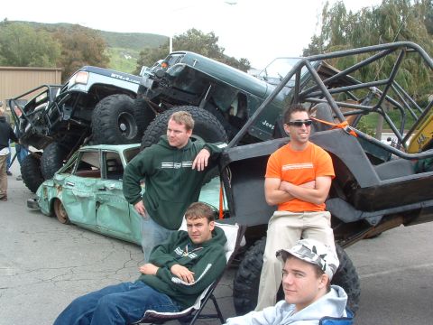 1994 Jeep YJ - Several heavily modified off-road vehicles are precariously stacked on top of a beat-up car, with onlookers gathered around.