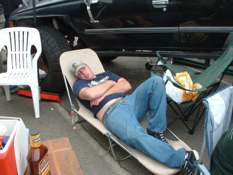 1994 Jeep YJ - Man naps in a lounge chair next to a lifted black vehicle, with camping chairs and a cooler visible.