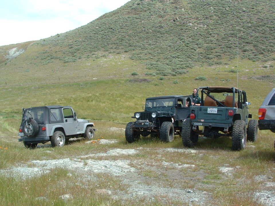 Three Jeeps parked in a grassy field with a hill in the background. One Jeep is silver, and two are dark green