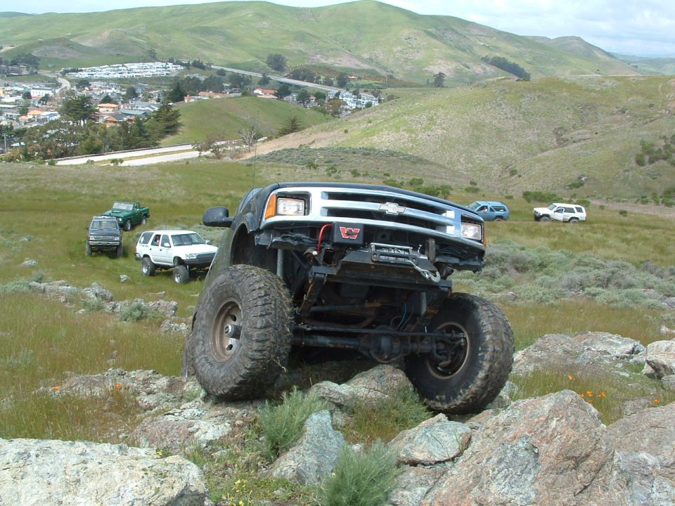 Modified Chevrolet pickup truck with large tires on rocky terrain, other SUVs and trucks in background