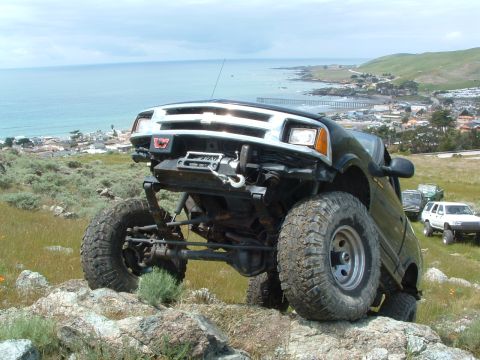 1994 Jeep YJ - This modified SUV is navigating a rocky trail with a coastal town and ocean in the distance.