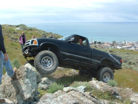 1994 Jeep YJ - A modified black pickup truck is shown off-roading on a steep, rocky incline with the ocean in the background.