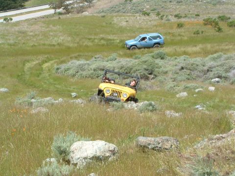 1994 Jeep YJ - Two off-road vehicles navigate a grassy, rocky terrain with a road visible in the background.