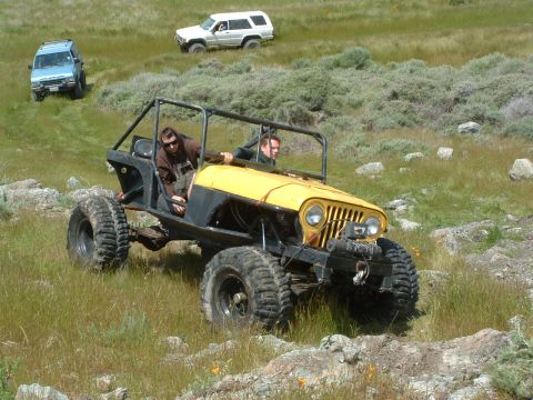 1994 Jeep YJ - A custom yellow buggy navigates a rocky, grassy terrain with other vehicles in the background.