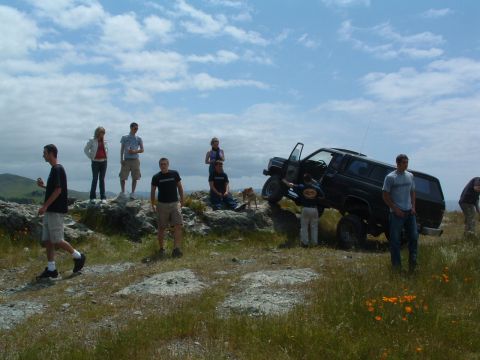 1994 Jeep YJ - Group of people gathered around a black SUV on a grassy, rocky hillside under a cloudy sky.