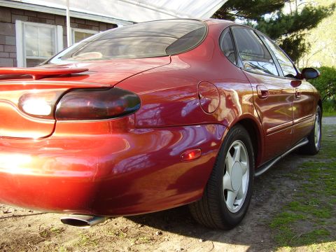 1997 Ford Taurus - The rear passenger side of a red sedan is visible, parked on a grassy area next to a building.