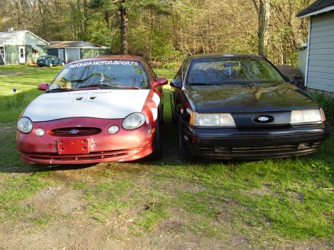 1997 Ford Taurus - Two Ford Taurus sedans are parked in a grassy area next to buildings.