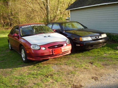 1997 Ford Taurus - Two Ford Taurus sedans, one red and one black, are parked side-by-side in a grassy area next to a building.