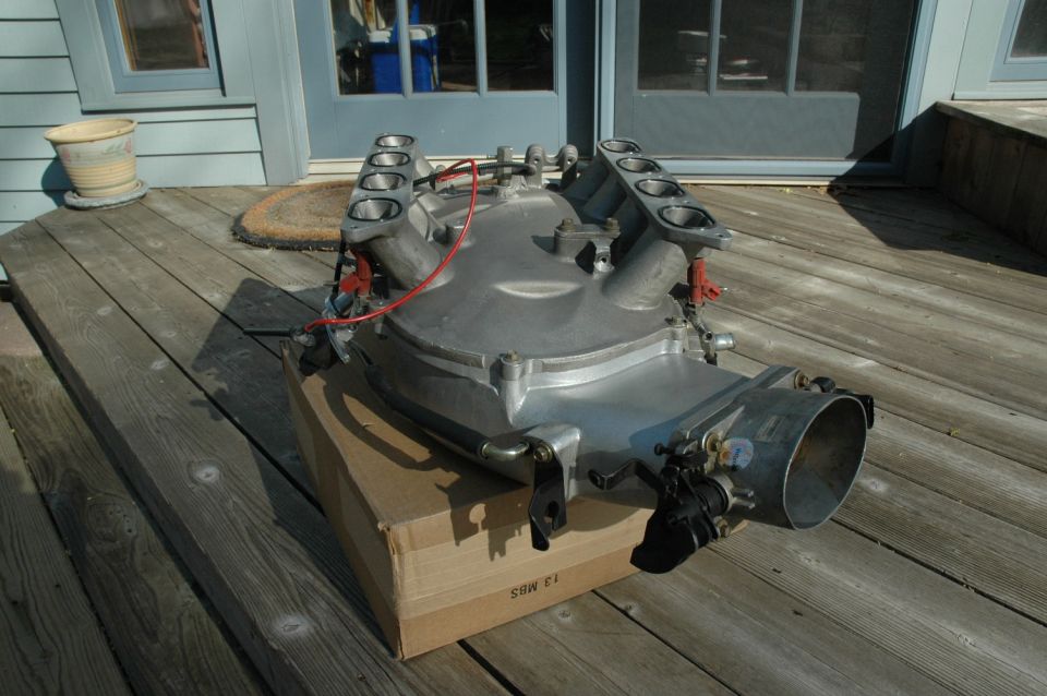 Aluminum intake manifold with eight intake ports and a throttle body, resting on a cardboard box on a wooden deck