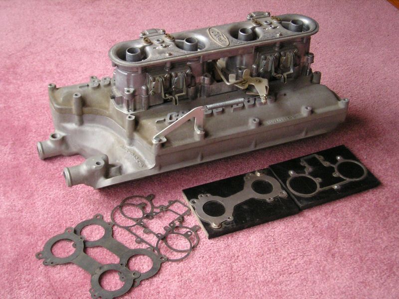 Aluminum Ford intake manifold with four carburetors and assorted gaskets on a pink surface