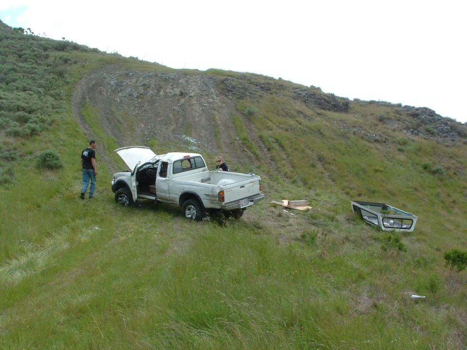 White pickup truck with hood open and door ajar on grassy hillside, removed truck cap nearby