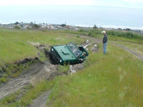1994 Jeep YJ - A green off-road vehicle has become stuck in a ditch on a grassy hillside, with a person observing the situation.