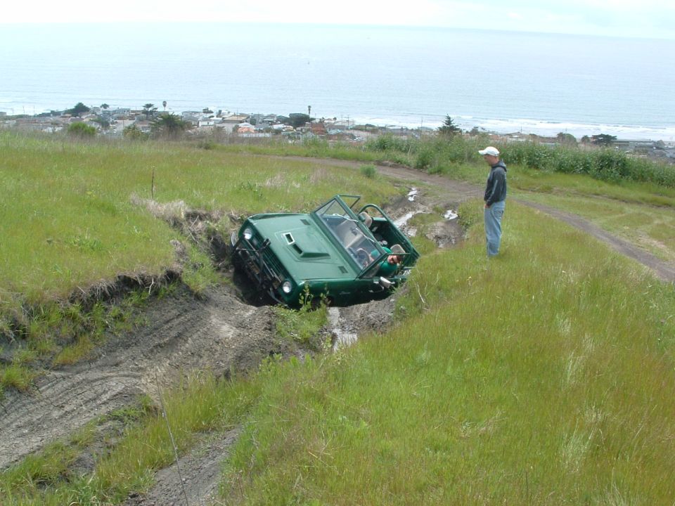 Green off-road vehicle tilted in a muddy ditch with a person standing nearby on a grassy hill overlooking the ocean