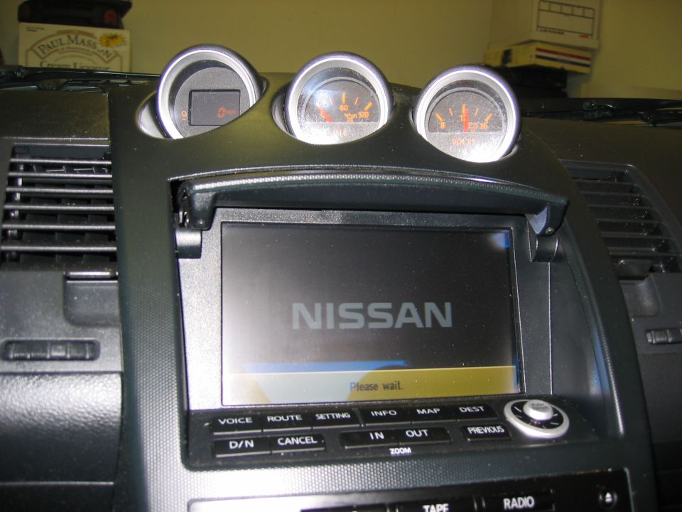 Nissan center dash with three hooded gauges above a nav screen showing the Nissan boot logo