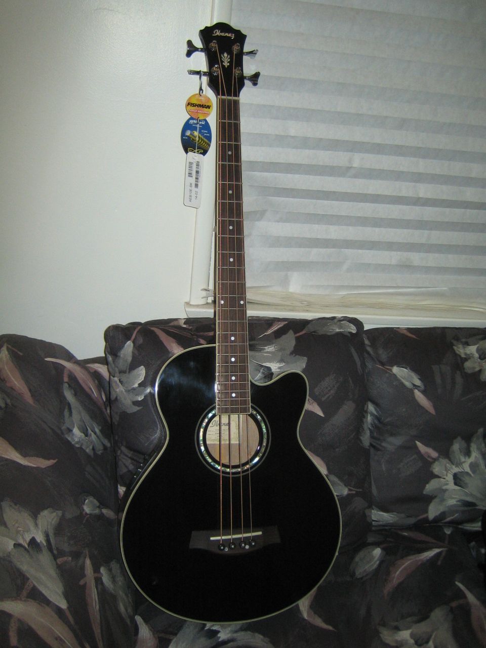 Black acoustic bass guitar with Ibanez logo on headstock, resting on a floral patterned couch