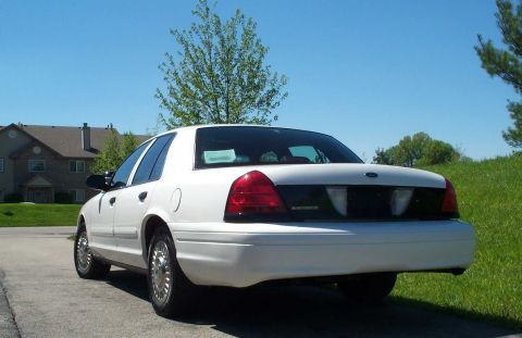 2001 Ford Crown Victoria - A white 2001 Ford Crown Victoria is parked on a paved surface with a grassy hill and houses in the background.