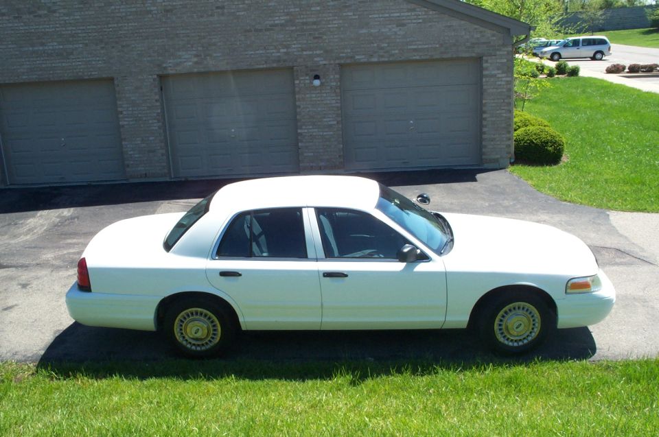 White Ford Crown Victoria sedan, side view, parked by brick garage with tan doors and green lawn