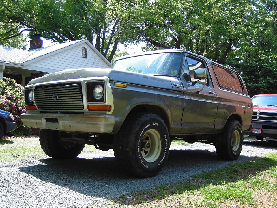 Faded green 1978-79 Ford Bronco with chrome grille and lift kit parked on a gravel driveway under trees